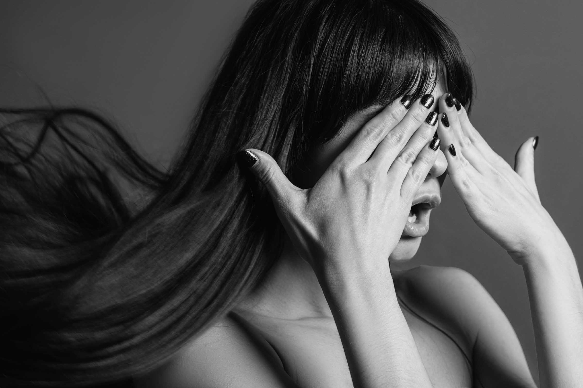 Black and white photo of a person with long hair covering their eyes with their hands. Their mouth is slightly open, and they have black painted nails. The person’s hair flows to the side, adding movement to the image—perfect inspiration for styles at the best hair salon Cary NC.